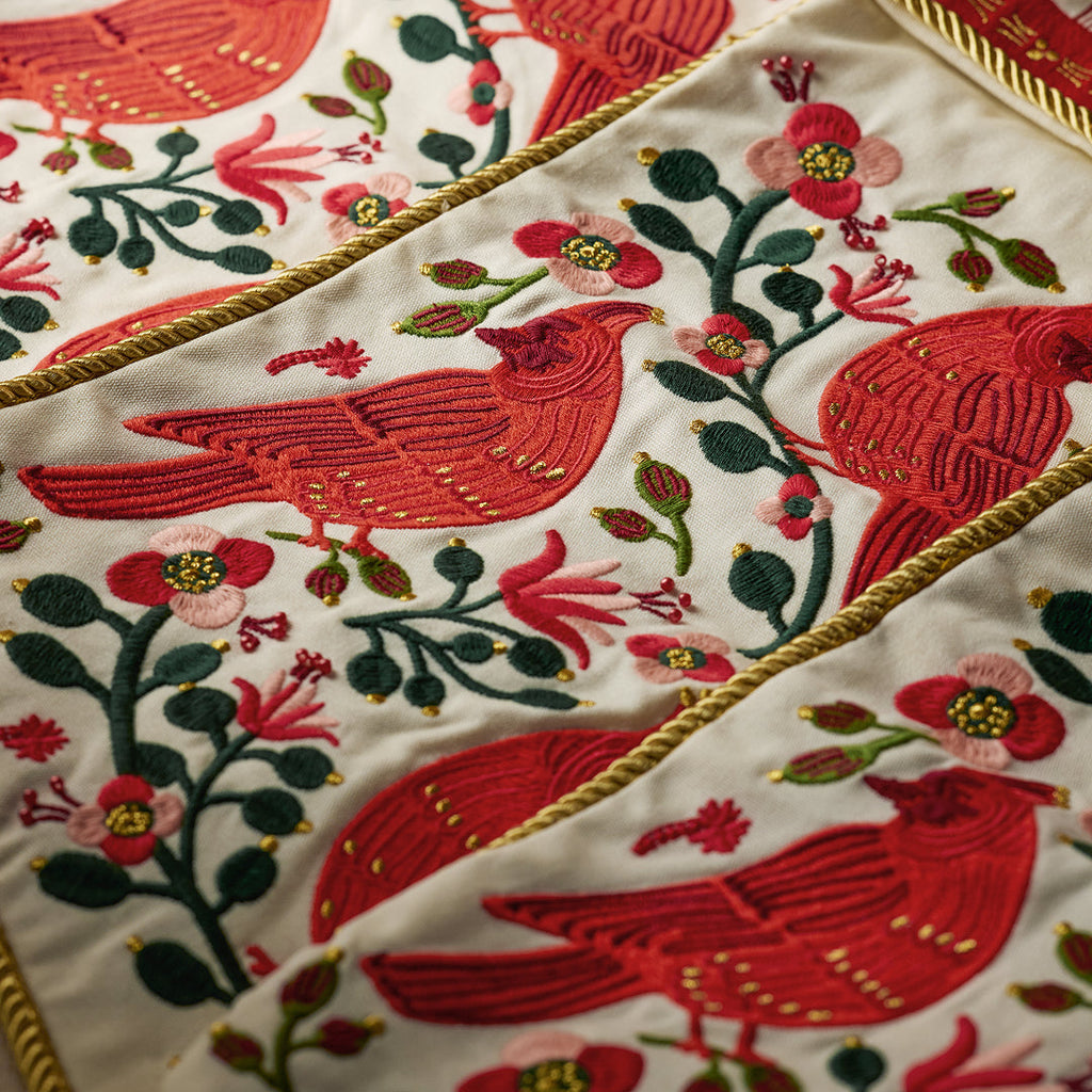 Close-up of a white embroidered textile featuring large red birds, green leaves, pink flowers, and gold decorative trim.