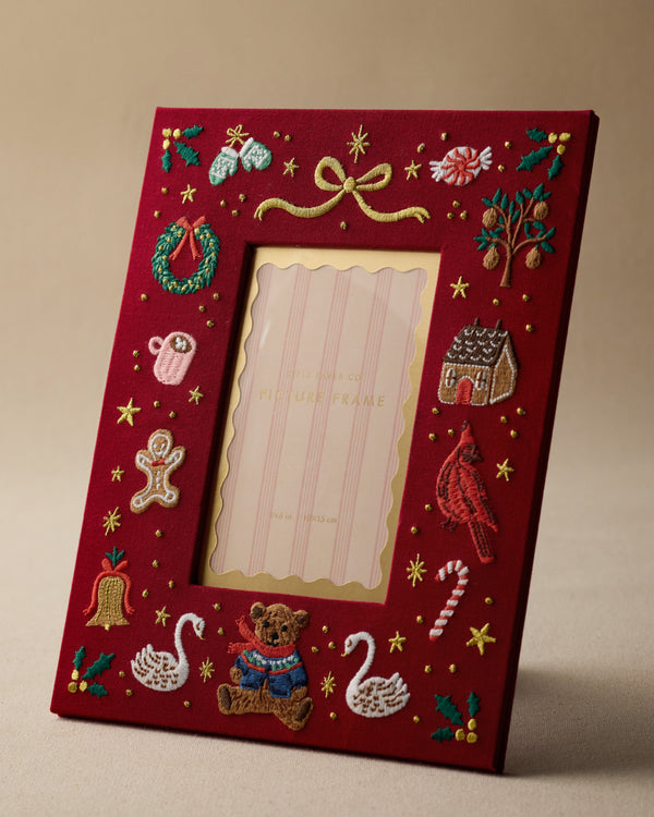 A red wooden picture frame with festive embroidered holiday designs, including a teddy bear, swans, candy cane, bird, wreath, and stars, shown on a neutral beige background.