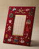 A red wooden picture frame with festive embroidered holiday designs, including a teddy bear, swans, candy cane, bird, wreath, and stars, shown on a neutral beige background.