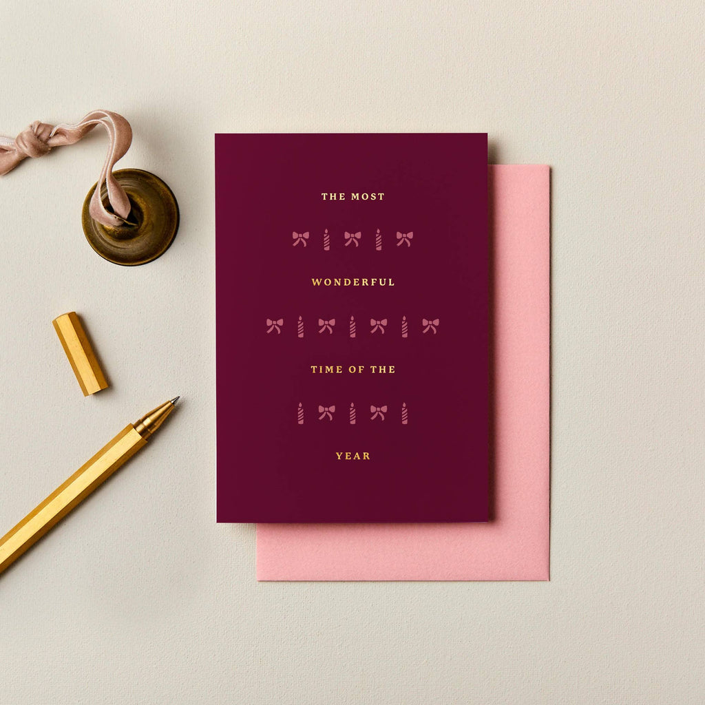 Burgundy A6 Christmas card with gold foil typography and festive icons, paired with a soft pink envelope.