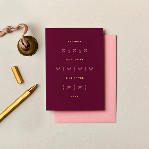 Burgundy A6 Christmas card with gold foil typography and festive icons, paired with a soft pink envelope.