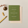 Olive green Christmas card with gold foil typography and festive icons, featuring a textured finish and cream envelope.