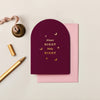 Burgundy arched Christmas card with gold foil moon and star motifs, paired with a pink envelope and gold pen.