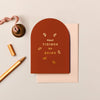 Rust-colored arched Christmas card with gold foil typography and botanical motifs on textured cardstock with an envelope.