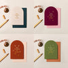 Four arched Christmas cards in cream, burgundy, rust, and olive with gold foil typography and textured card stock.