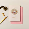 Ink & Bloom Christmas card with a festive wreath illustration, gold foil lettering, and a pink envelope on textured paper.