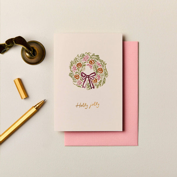 Ink & Bloom Christmas card with a festive wreath illustration, gold foil lettering, and a pink envelope on textured paper.