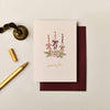Textured cream Christmas card with candle illustration, gold foil lettering, and deep purple envelope on a desk.