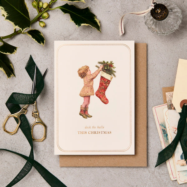 Christmas card with a child and stocking, surrounded by festive items on a textured surface.