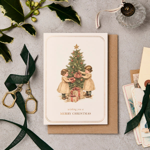 Christmas card with two children around a tree, surrounded by festive decorations on a gray surface.