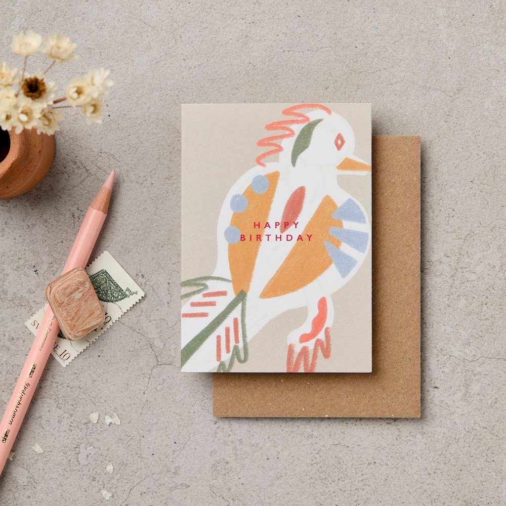 Greeting card with a colorful bird illustration and 'Happy Birthday' text, placed on a textured surface with a pen and small plant.