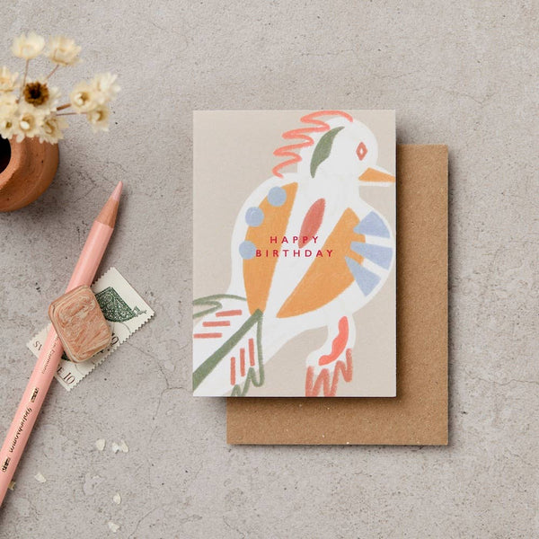 Greeting card with a colorful bird illustration and 'Happy Birthday' text, placed on a textured surface with a pen and small plant.