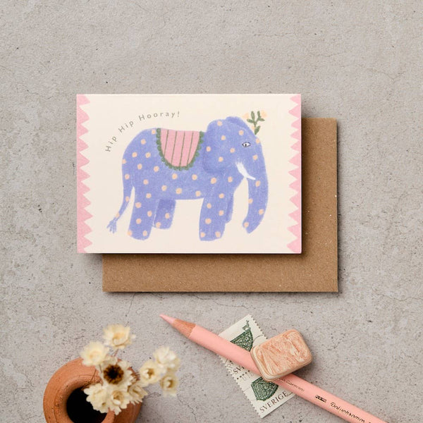 A greeting card featuring a whimsical blue elephant with pink polka dots and a pink patterned saddle, placed on top of a brown kraft envelope. The card is styled on a textured gray surface with a pink pencil, a small ceramic pot with dried flowers, and a stamp.