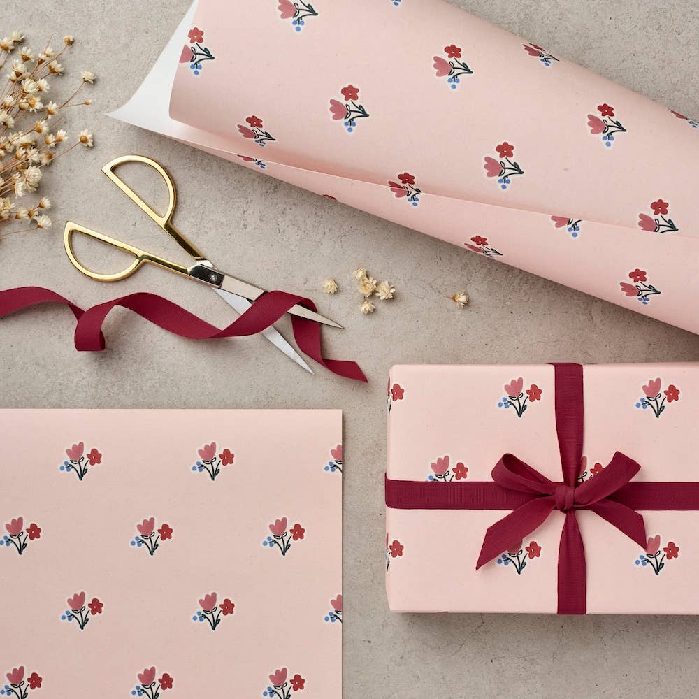 Pink floral wrapping paper shown rolled out and flat, with a gift box wrapped in matching paper and tied with a burgundy ribbon. Gold-handled scissors and dried flowers are arranged on a light gray surface.