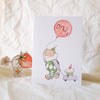 Greeting card with a cartoon character, balloon, and cupcake on a white surface.