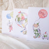 Three children's greeting cards with illustrations of a clown, hot air balloon, and balloons on a textured surface.