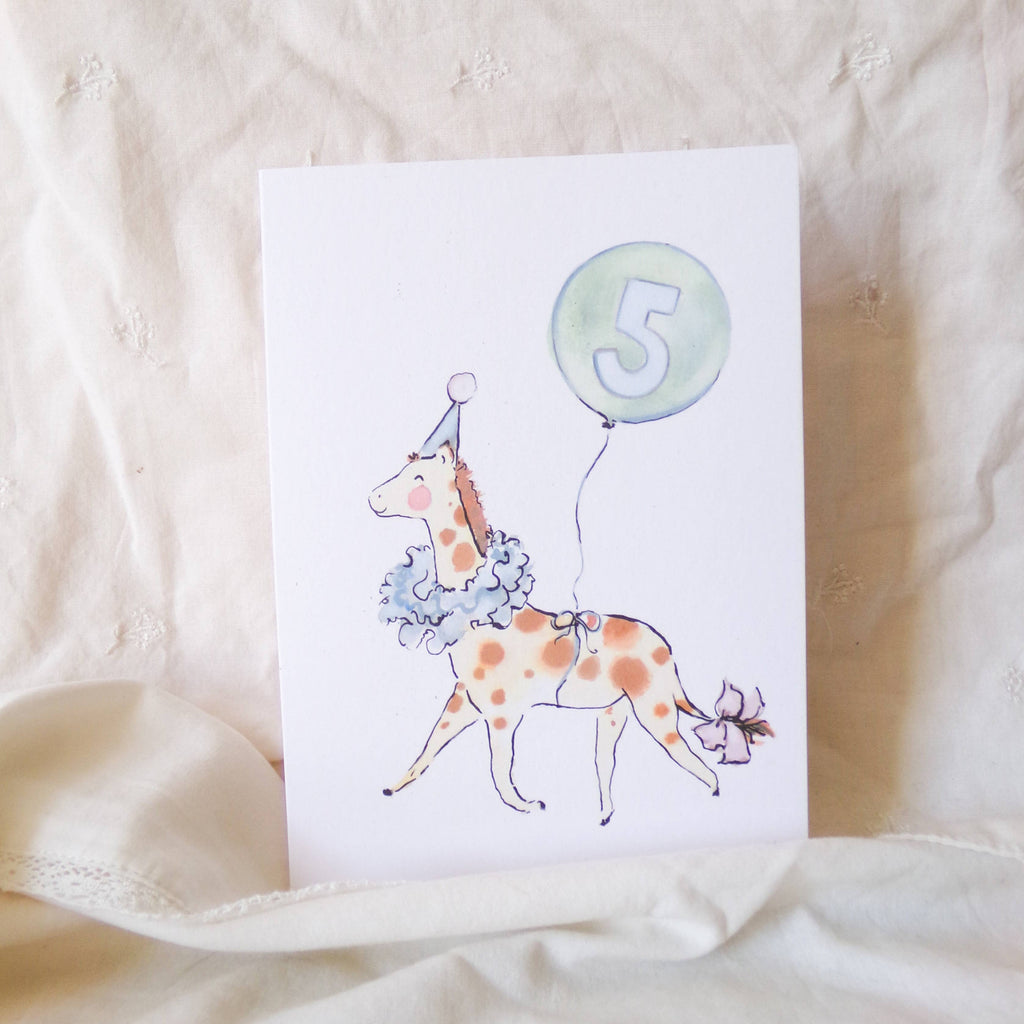 Card with a giraffe illustration and number 5 balloon on a beige fabric background