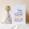Children's birthday hat and card with cake illustration on a white surface