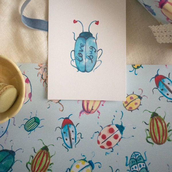 Note card with a beetle design on a table with colorful beetle-patterned paper and a bowl.