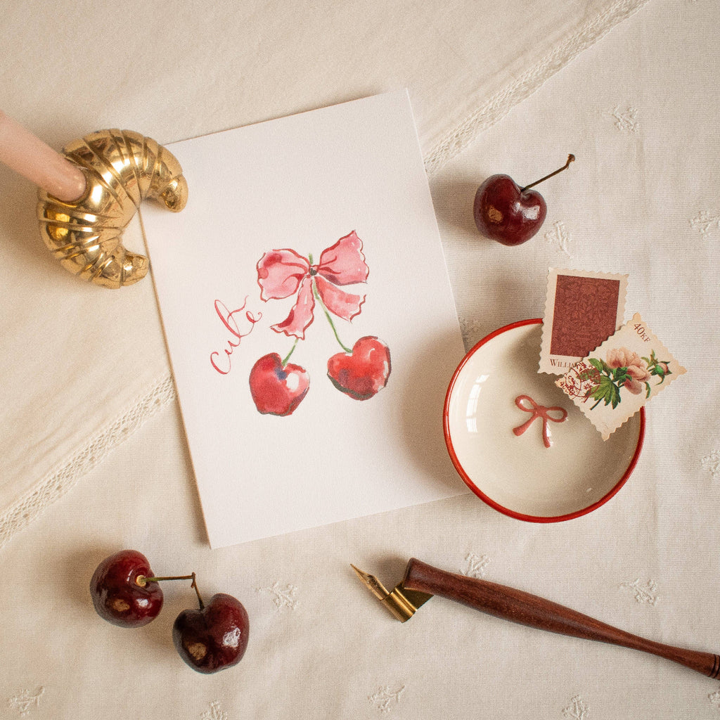 Decorative card with cherries and a bow on a textured surface with cherries and a pen.