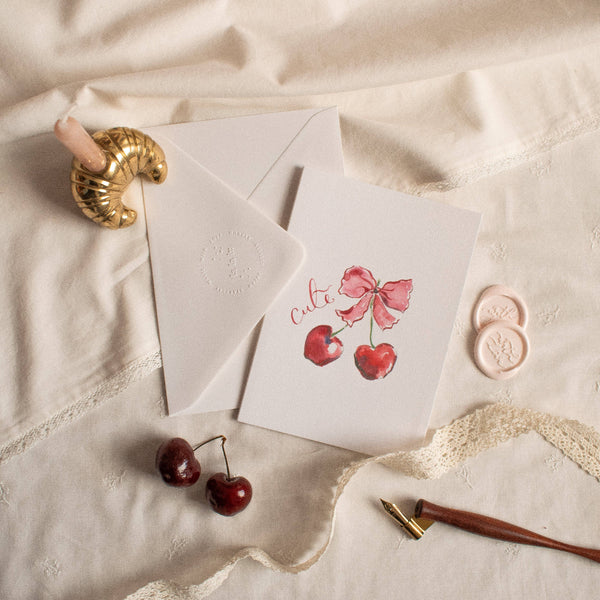 Card with cherries and a bow on a textured surface with a gold ring and red cherries.