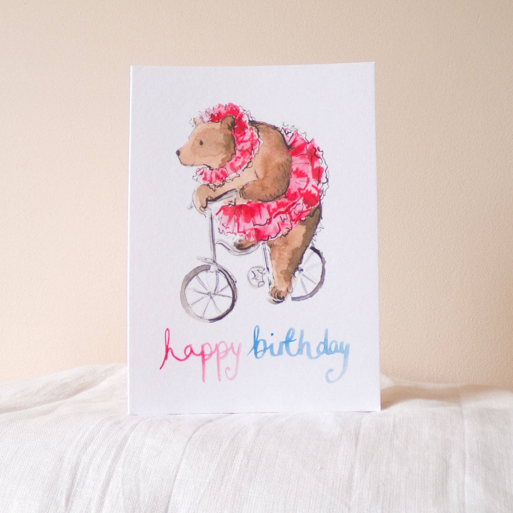Greeting card with a bear riding a bike and wearing a pink outfit, on a white surface.