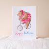 Greeting card with a bear riding a bike and wearing a pink outfit, on a white surface.