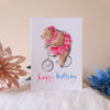 Greeting card with a bear riding a bicycle and 'happy birthday' text, placed on a surface with decorative flowers.