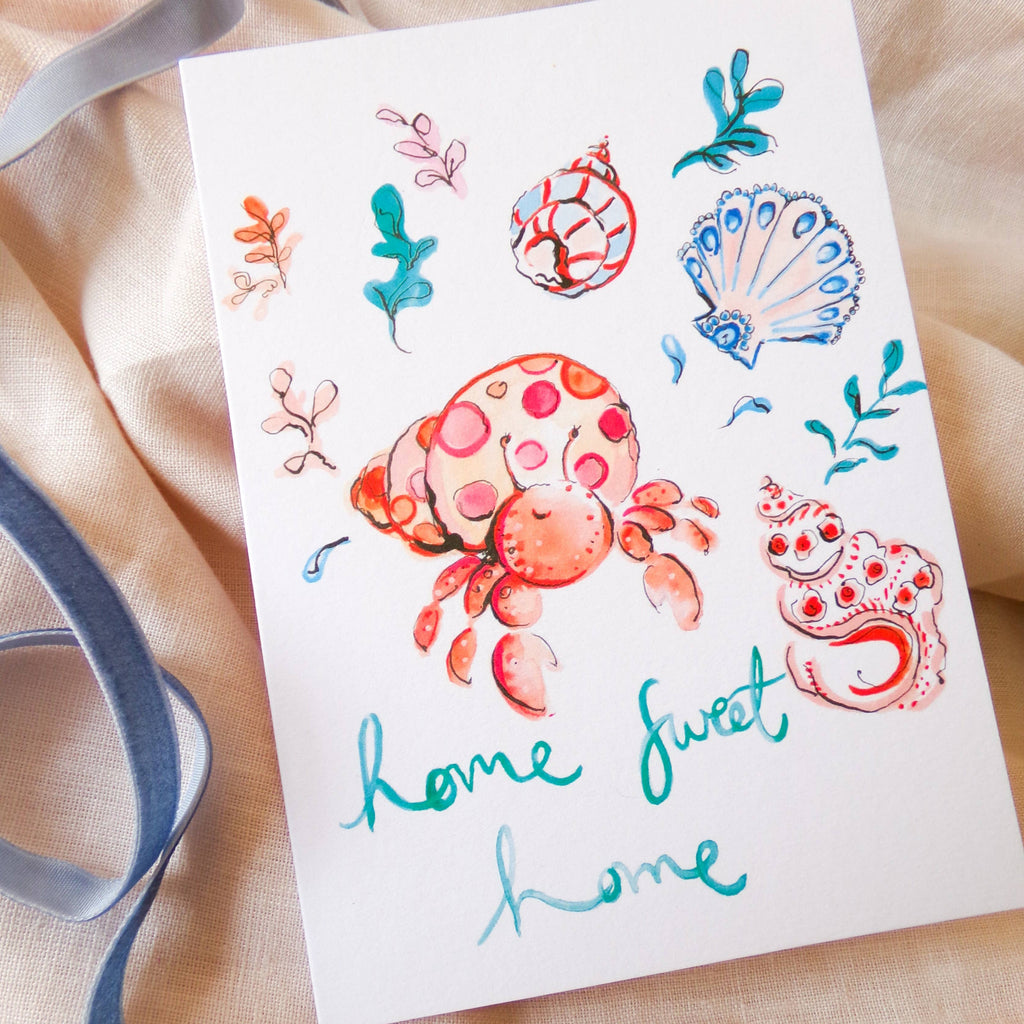 Colorful tattoo designs of crabs and shells on a white card with 'home sweet home' text, placed on a beige fabric background.