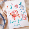 Colorful tattoo designs of crabs and shells on a white card with 'home sweet home' text, placed on a beige fabric background.