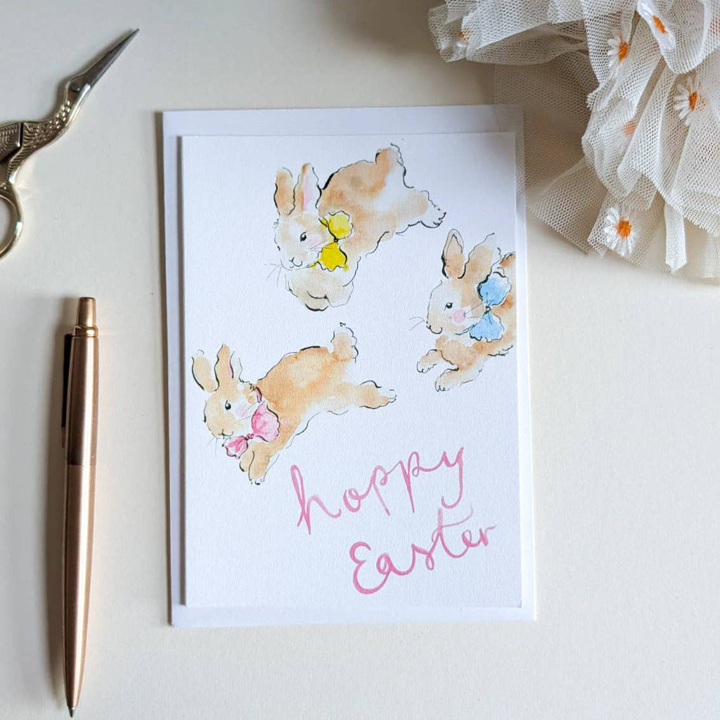 Easter card with bunnies and 'Happy Easter' text on a white surface with a pen and decorative bow.