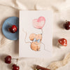 Card with a mouse holding a heart-shaped balloon on a textured surface with cherries and a pen.