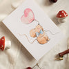 Card with teddy bear and heart balloon illustration on a textured surface with mushrooms and cherries.