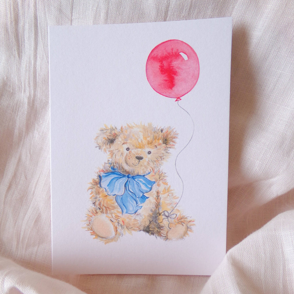 Card with a teddy bear holding a red balloon on a soft fabric background