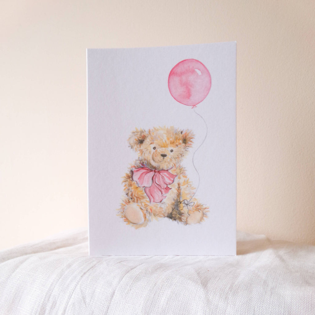 Card with a teddy bear holding a pink balloon on a white surface.