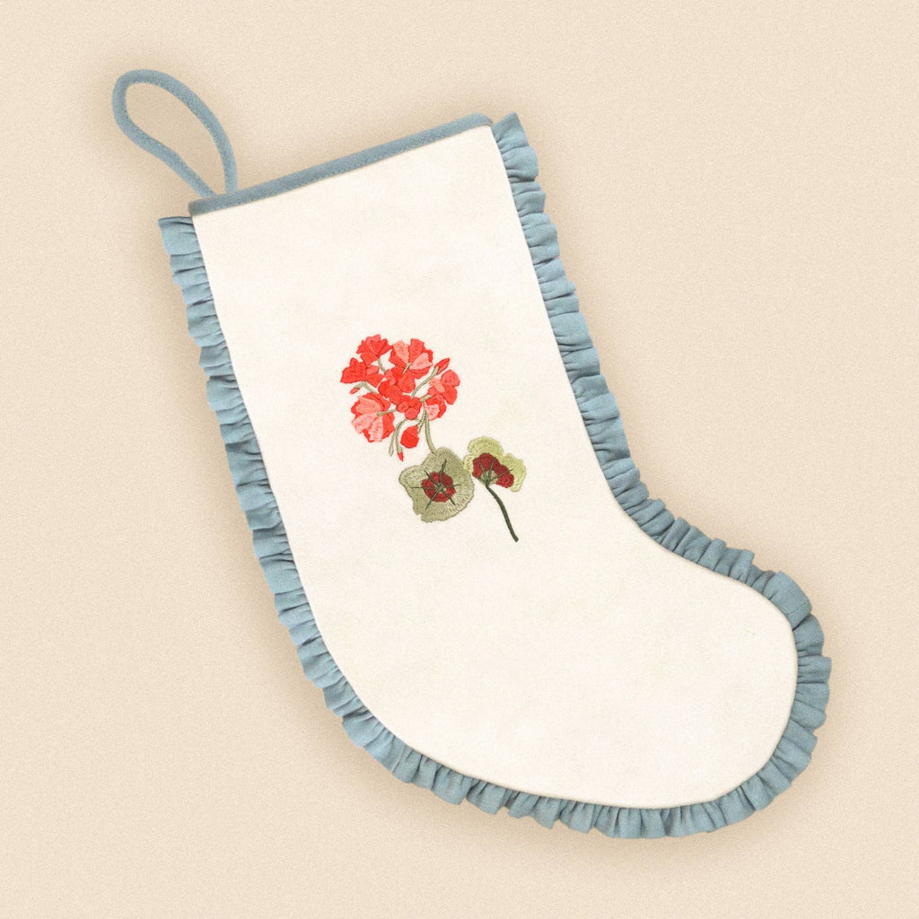 An off-white fabric stocking with a light blue ruffled trim and a hanging loop, featuring an embroidered red flower and green leaves on the front, set against a solid beige background.