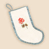 An off-white fabric stocking with a light blue ruffled trim and a hanging loop, featuring an embroidered red flower and green leaves on the front, set against a solid beige background.