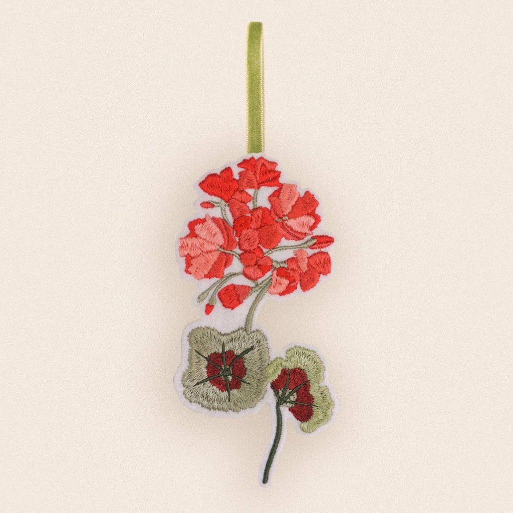An embroidered fabric ornament featuring red geranium flowers and green leaves hanging from a light green ribbon on a plain cream background.