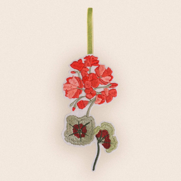 An embroidered fabric ornament featuring red geranium flowers and green leaves hanging from a light green ribbon on a plain cream background.