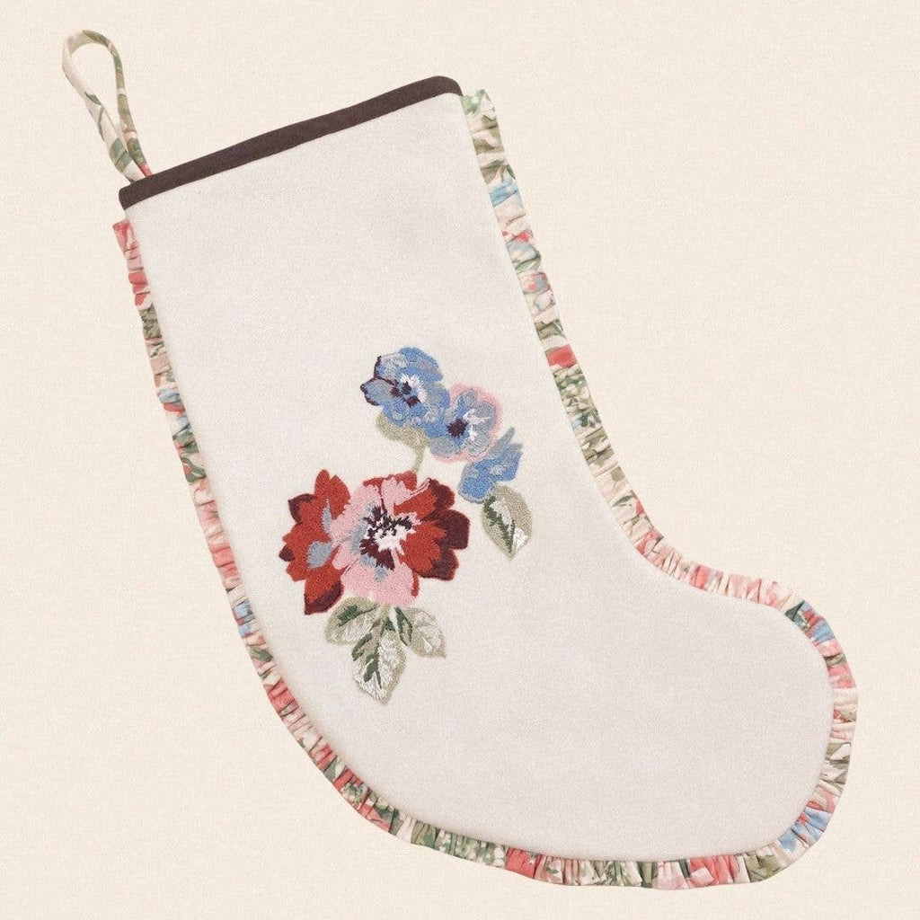 A white embroidered Christmas stocking with a floral design in red, pink, and blue, featuring a multicolor patterned trim and a hanging loop, shown on a light beige background.