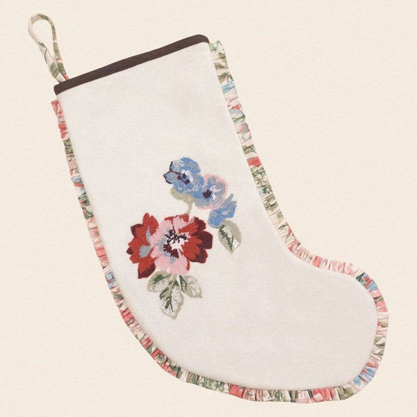 A white embroidered Christmas stocking with a floral design in red, pink, and blue, featuring a multicolor patterned trim and a hanging loop, shown on a light beige background.