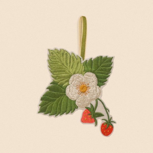 An embroidered strawberry ornament featuring a white flower, green leaves, and two red strawberries on a beige ribbon, shown against a solid cream background.