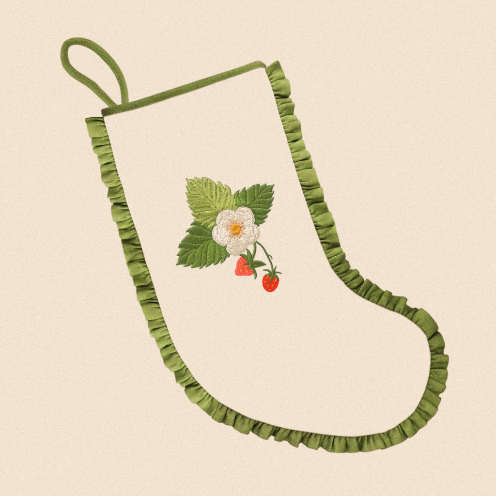 A cream-colored fabric stocking with a green ruffled trim and a hanging loop, featuring an embroidered strawberry and white flower motif in the center, set against a solid beige background.