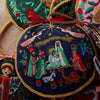 Embroidered nativity scene ornament with gold trim on a textured surface.