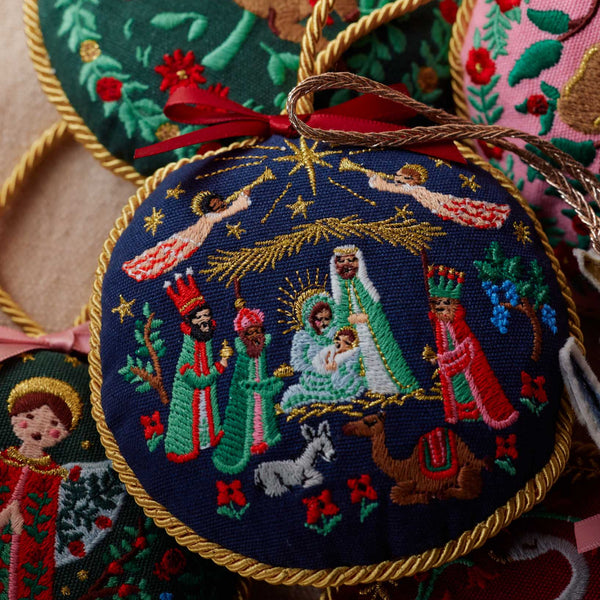 Embroidered nativity scene ornament with gold trim on a textured surface.