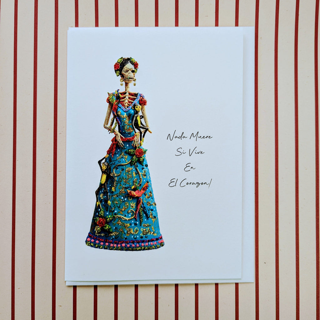 A greeting card with an illustration of a skeleton wearing a traditional, colorful outfit, positioned against a striped background. The card has a title and signature-like text at the bottom.
