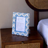 Decorative frame with a blue and white pattern on a wooden nightstand next to a bed with a wicker headboard.