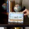 Hand holding a photo card with a beach scene on a wooden surface with books and globes in the background.