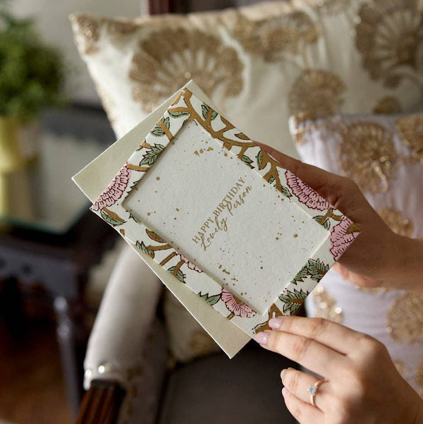 Hand holding a decorative card with floral designs in a cozy indoor setting.