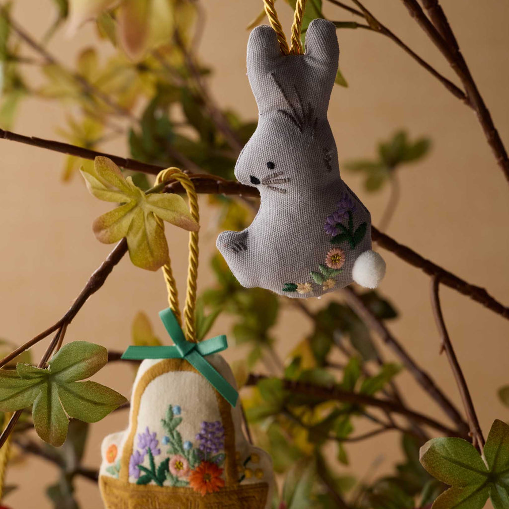 Decorative bunny and basket ornament hanging on a branch with leaves.